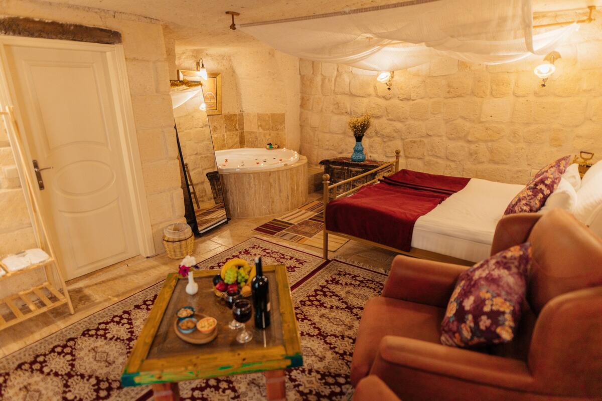 A cozy room with a bed, a small table with a vase and candles, a stone wall, and a wooden door.
