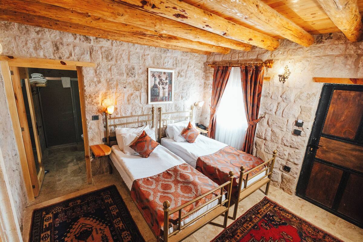 A rustic room with two beds, a wooden ceiling, stone walls, and a door.