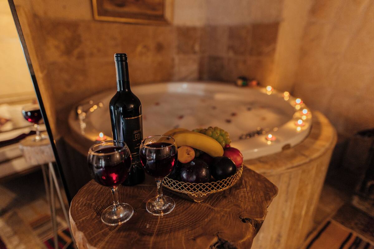 A wooden table with a bottle of wine, two wine glasses, and a fruit basket. In the background, there is a hot tub with candles around it.