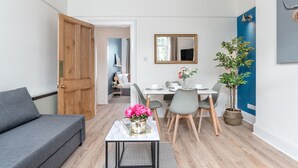 A modern living room with a grey sofa, a small table with a vase of flowers, a dining area with a white table and grey chairs, and a blue accent wall. - West Mount Apartment (Aberdeen)