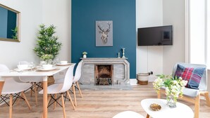 A modern living room with a fireplace, a dining table with chairs, a TV mounted on the wall, and a sofa with a plaid cushion. - West Mount Apartment (Aberdeen)