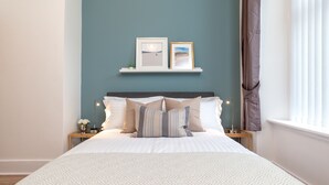 A neatly made bed with patterned bedding, a bedside table with a lamp, and a wall-mounted shelf with two framed pictures. - West Mount Apartment (Aberdeen)