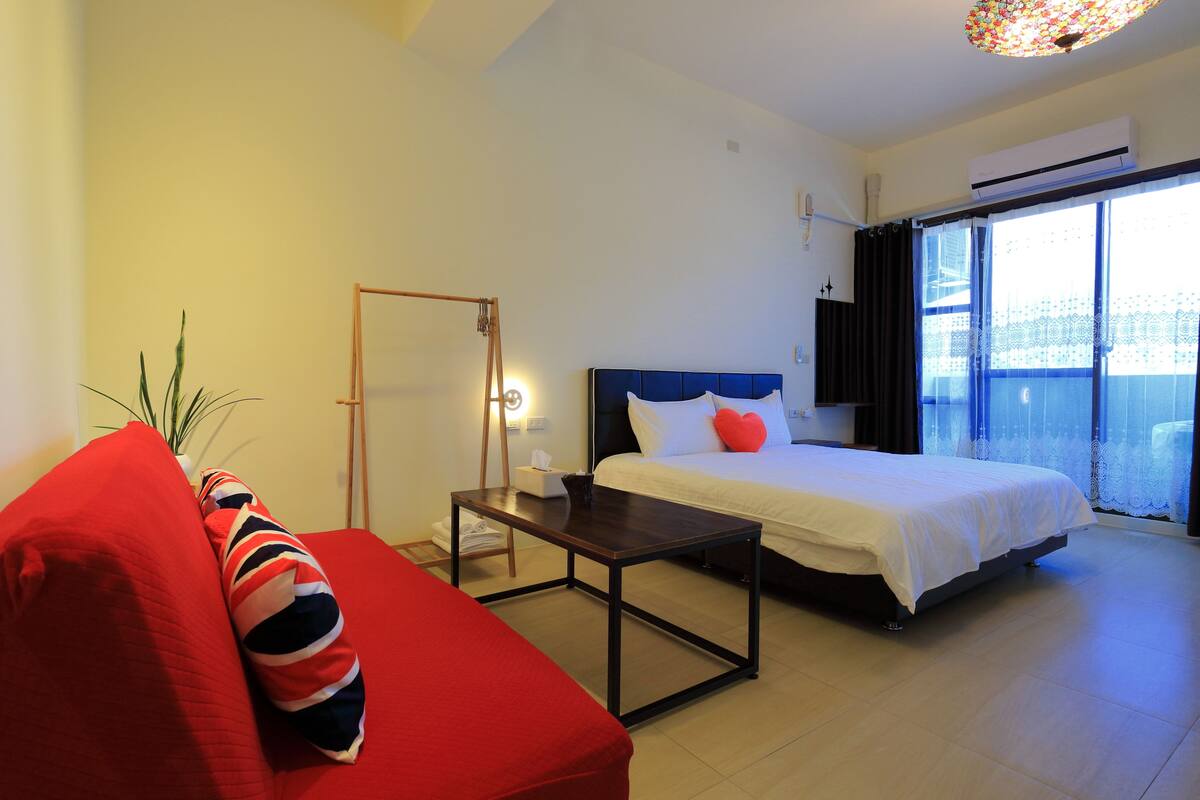 Una habitación de hotel moderna con una cama, un sofá rojo, una mesita y un ventanal con cortinas.