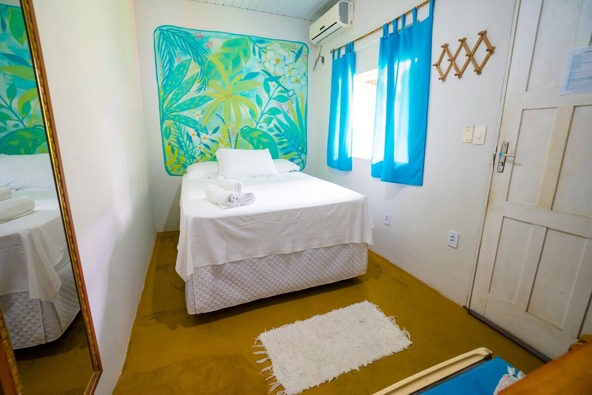 A bedroom with a bed, a mirror, a tropical-themed wall, a window with blue curtains, and a door.
