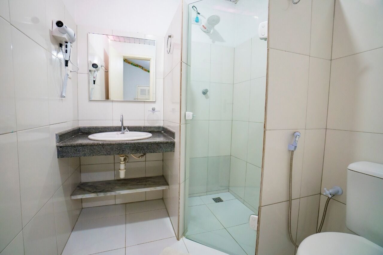 A bathroom with a granite countertop sink, a mirror, a hairdryer mounted on the wall, and a glass-enclosed shower area.