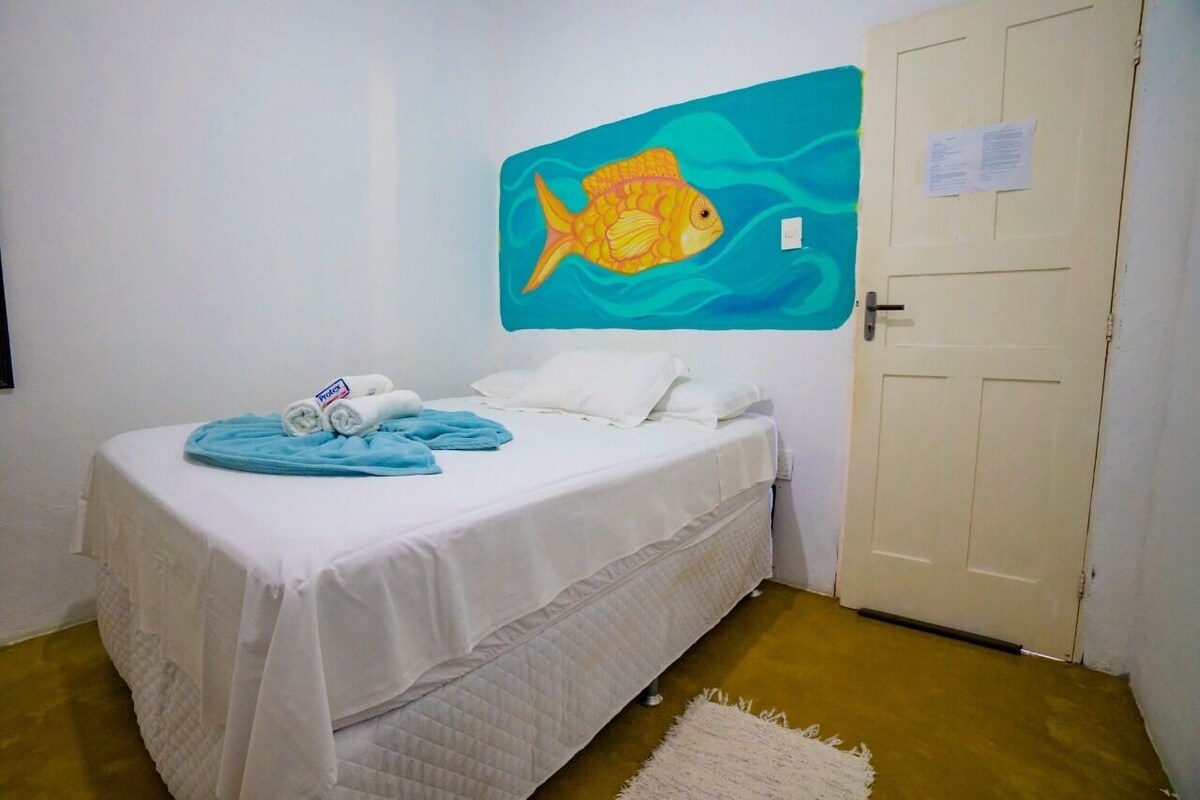 A single bed with white bedding, a blue blanket, and rolled towels. A painting of a fish is on the wall.