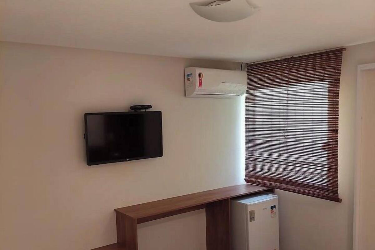 A hotel room with a bed, a TV mounted on the wall, a small air conditioning unit, and a window with blinds.