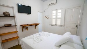 A hotel room with a bed, a television, a window with shutters, and a shelf with decorative items. - Pousada Sunset Boipeba (Cairu)