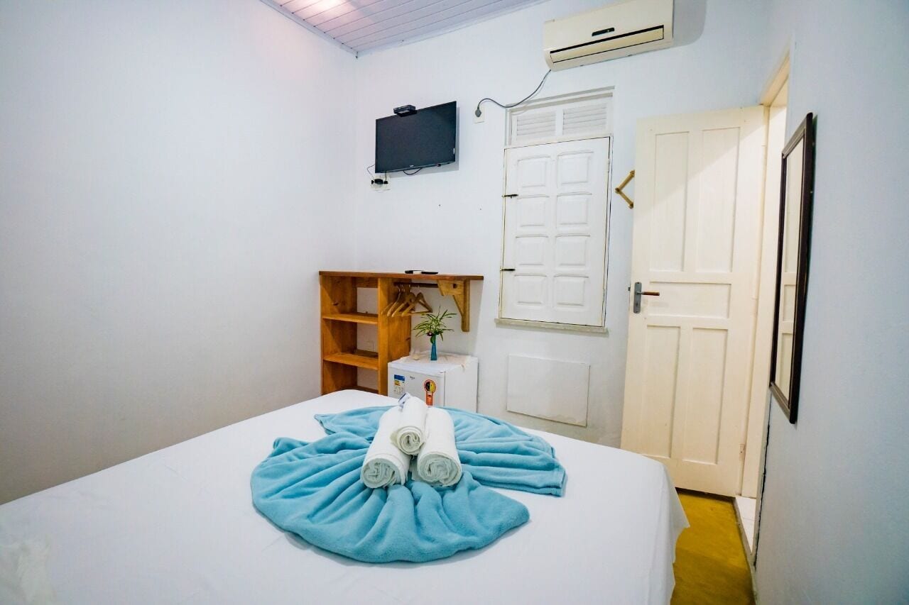 A hotel room with a bed, a TV mounted on the wall, an air conditioning unit, a wooden shelf with a plant, and a door.