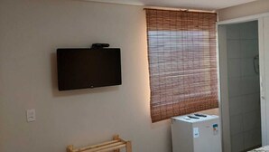 A small, clean room with a TV mounted on the wall, a wooden shelf, a window with blinds, and a bed with a floral bedspread. - Pousada Sunset Boipeba (Cairu)