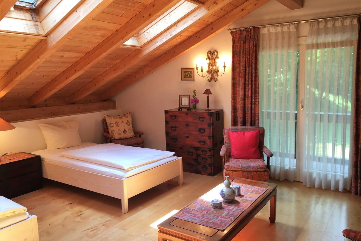 A cozy attic bedroom with a wooden ceiling, a bed, a dresser, a chair, and a table with decorative items.