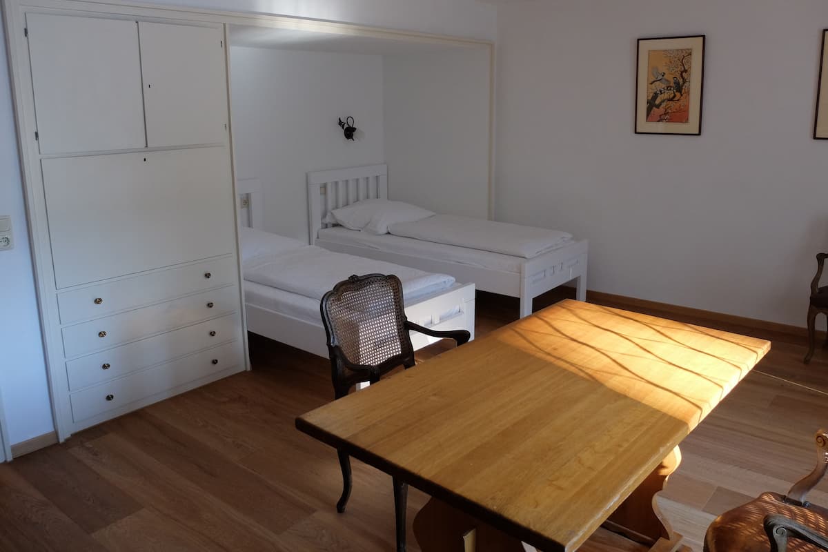 A room with a wooden floor, a white bed with a headboard, a wooden table, a chair, and a wardrobe with drawers.