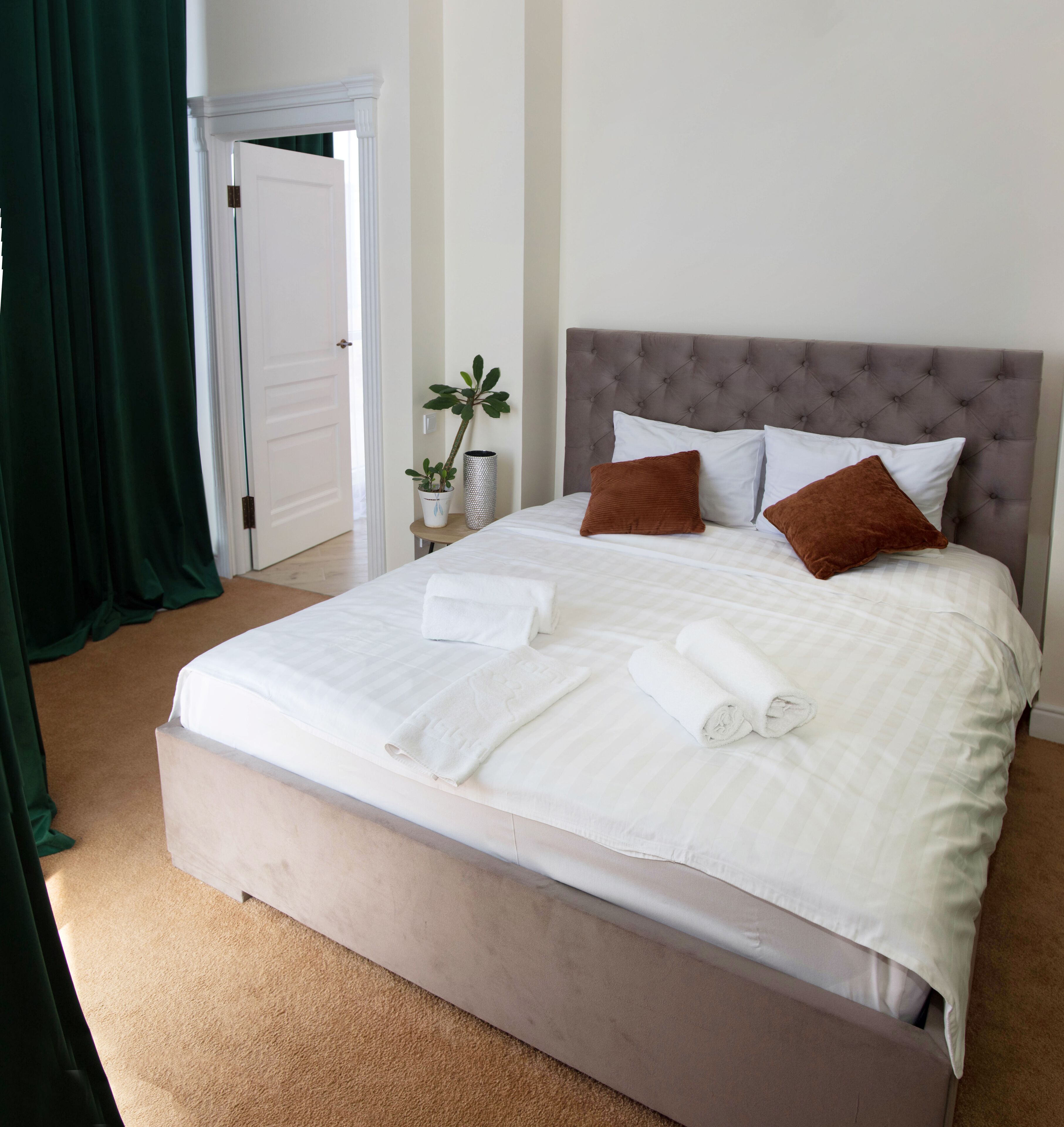 A neatly made bed with white linens and brown pillows, a small potted plant, and a white door in the background.