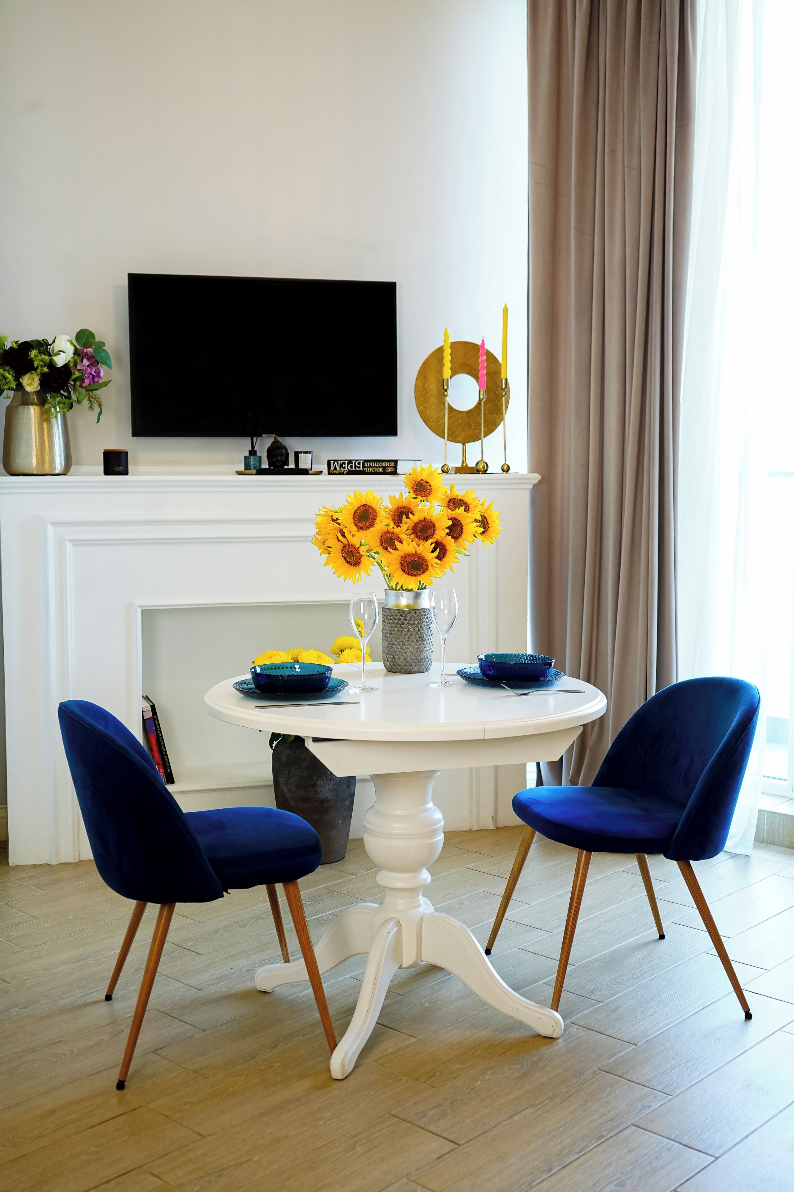 Een eetruimte met een witte tafel, blauwe stoelen, een vaas met zonnebloemen en een flatscreen televisie boven een open haard.