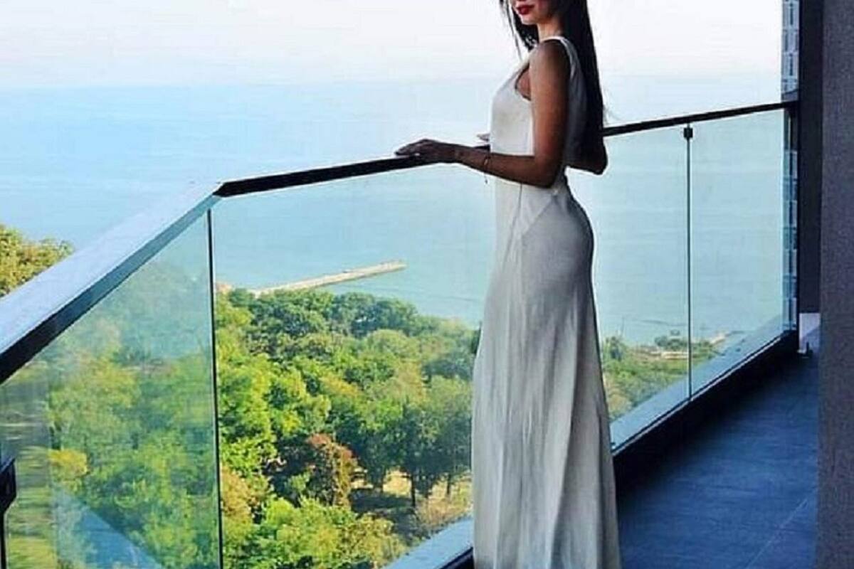 Eine Frau im weißen Kleid steht auf einem Balkon mit Blick auf einen Wald und das Meer.
