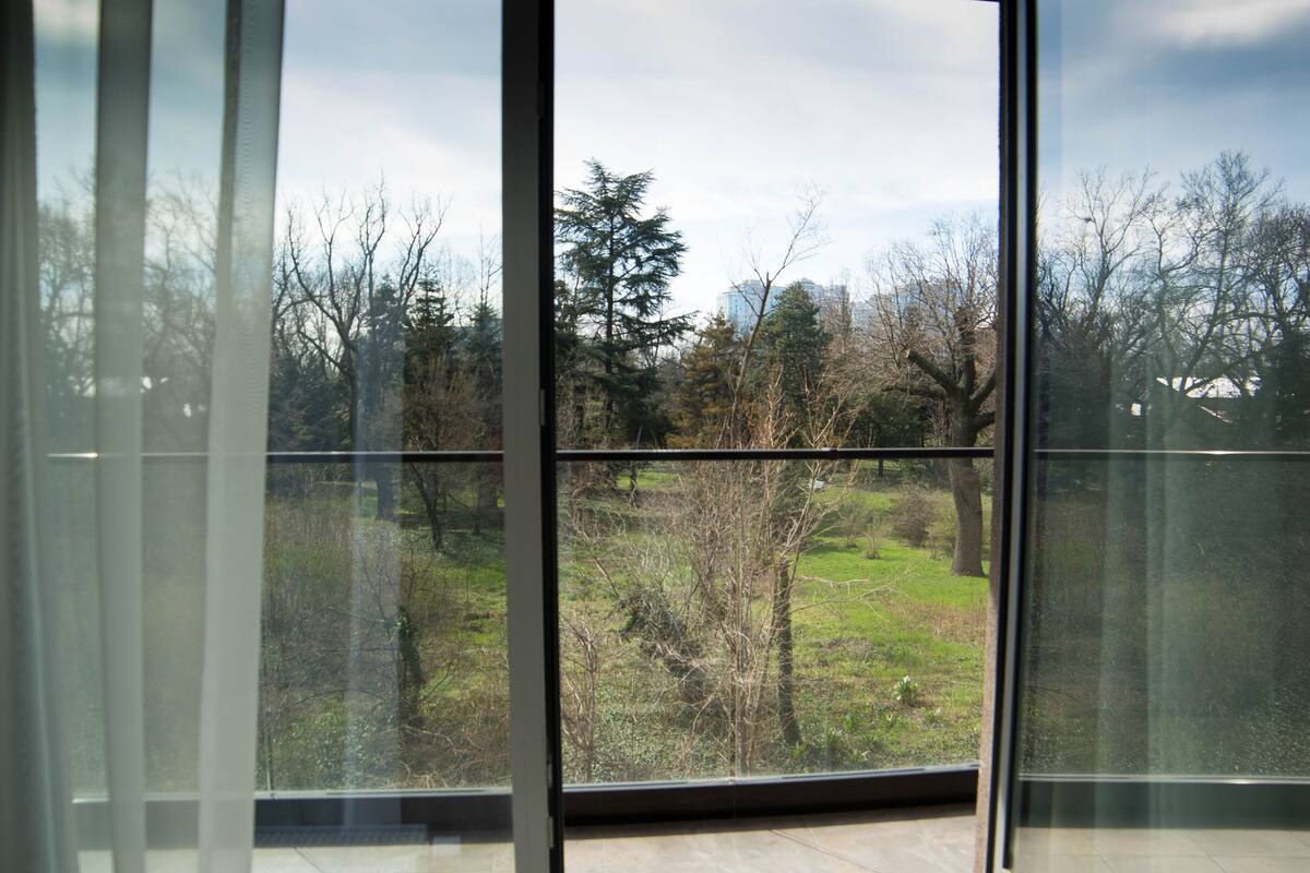 Ein Zimmer mit großen Fenstern, das einen Blick auf eine Grünfläche mit Bäumen und ein Gebäude in der Ferne bietet.