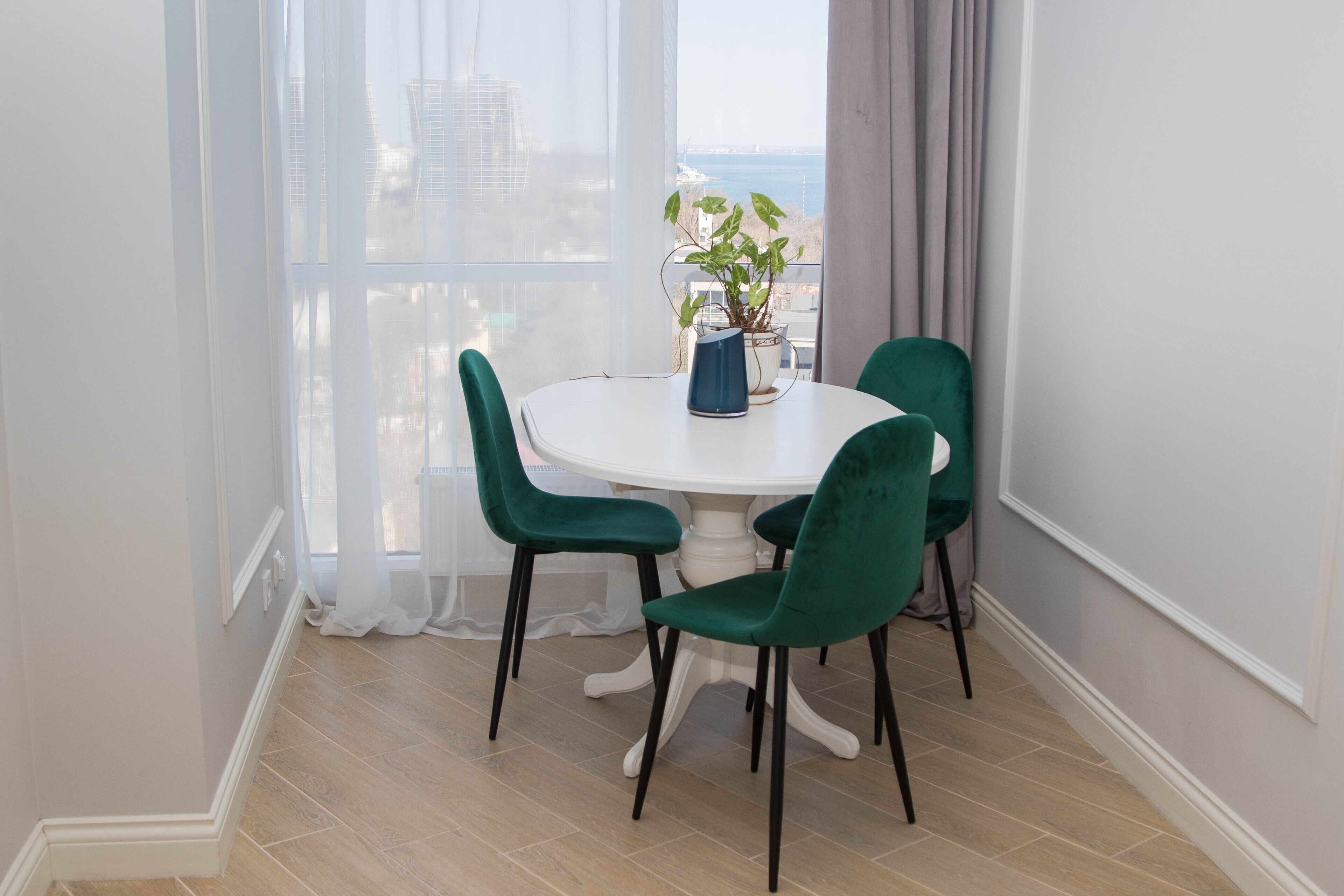Un coin repas avec une table ronde, quatre chaises vertes, une plante en pot et une vue sur la ville grâce à de grandes fenêtres.