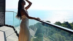 Une femme, vêtue d’une longue robe, se tient près d’une rambarde en verre, d’où l’on peut admirer l’océan et la végétation en contrebas.