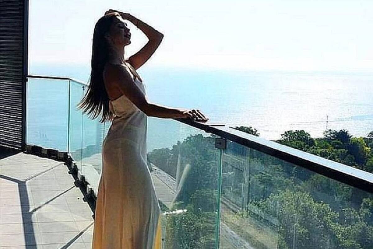 Eine Frau in einem langen Kleid steht an einer Glasuhr und blickt auf das Meer und das darunterliegende Grün.