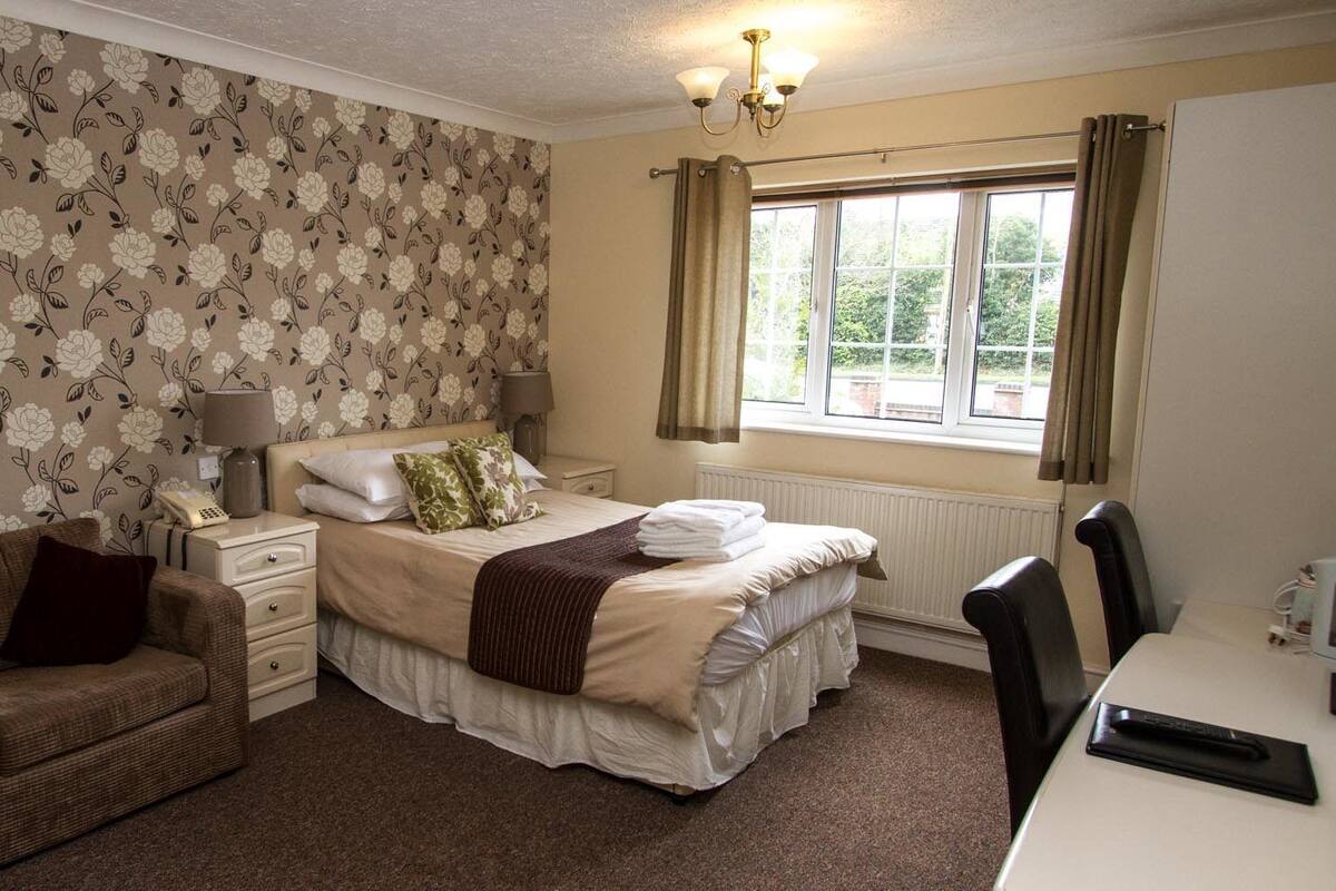 A bedroom with a floral wallpaper, a bed with pillows, a bedside table with a lamp, a window with curtains, and a chair.
