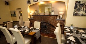 Un comedor con chimenea de ladrillo, mesas dispuestas con platos y cubiertos, y sillas.