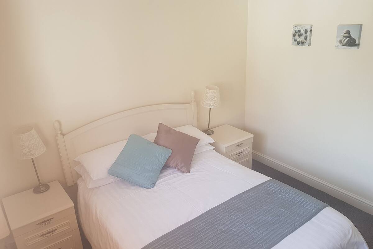 A neatly made bed with a grey quilt, two pillows, a bedside table with a lamp, and a small drawer unit.
