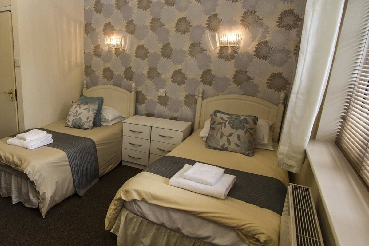 A hotel room with two single beds, a desk with a chair, a small table with a kettle, and a door with a notice.