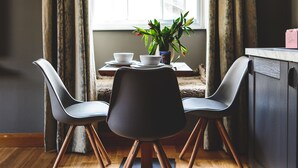 Uma área de refeições com uma mesa, duas cadeiras, uma planta em vaso e uma janela com cortinas.