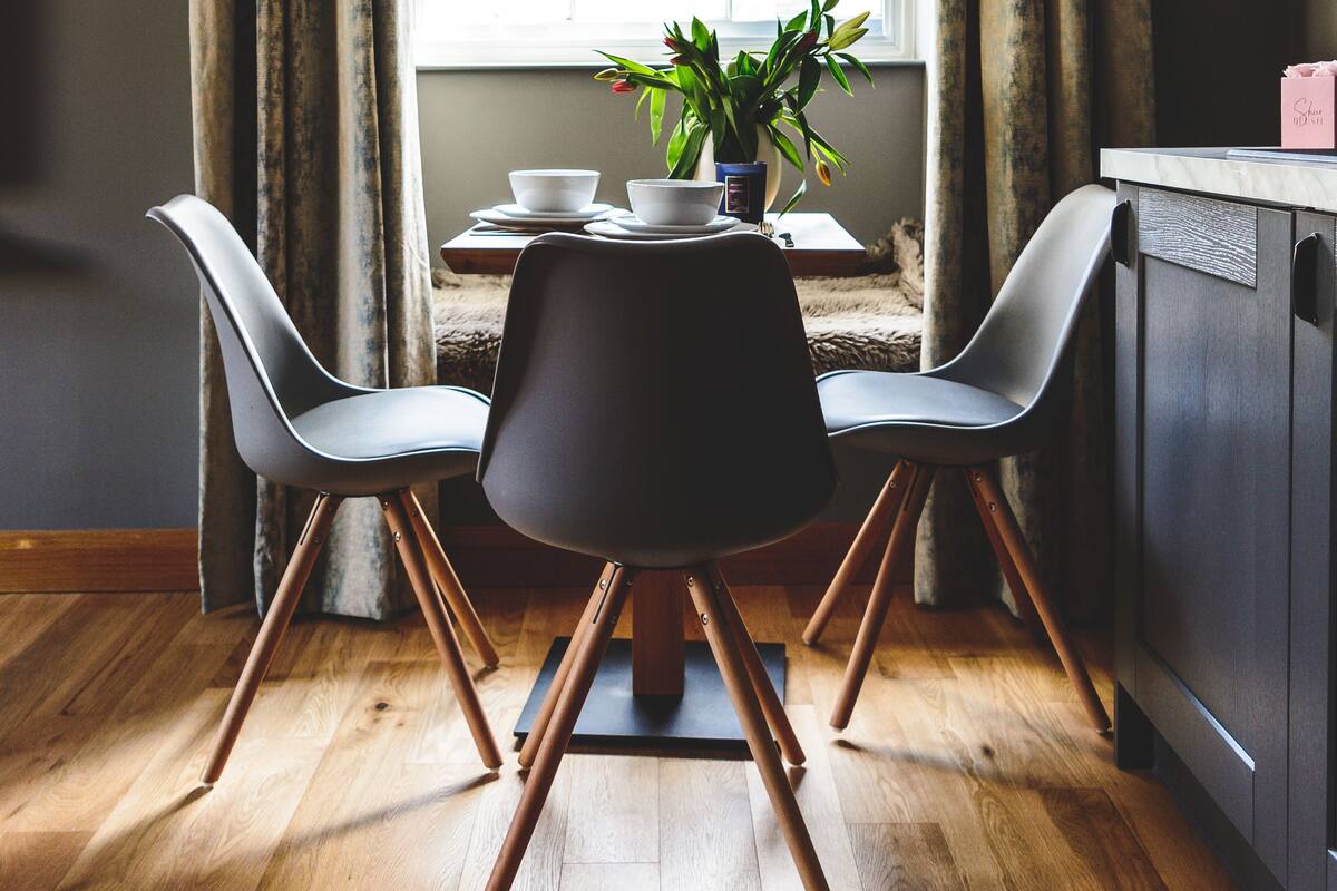 Uma área de refeições com uma mesa, duas cadeiras, uma planta em vaso e uma janela com cortinas.