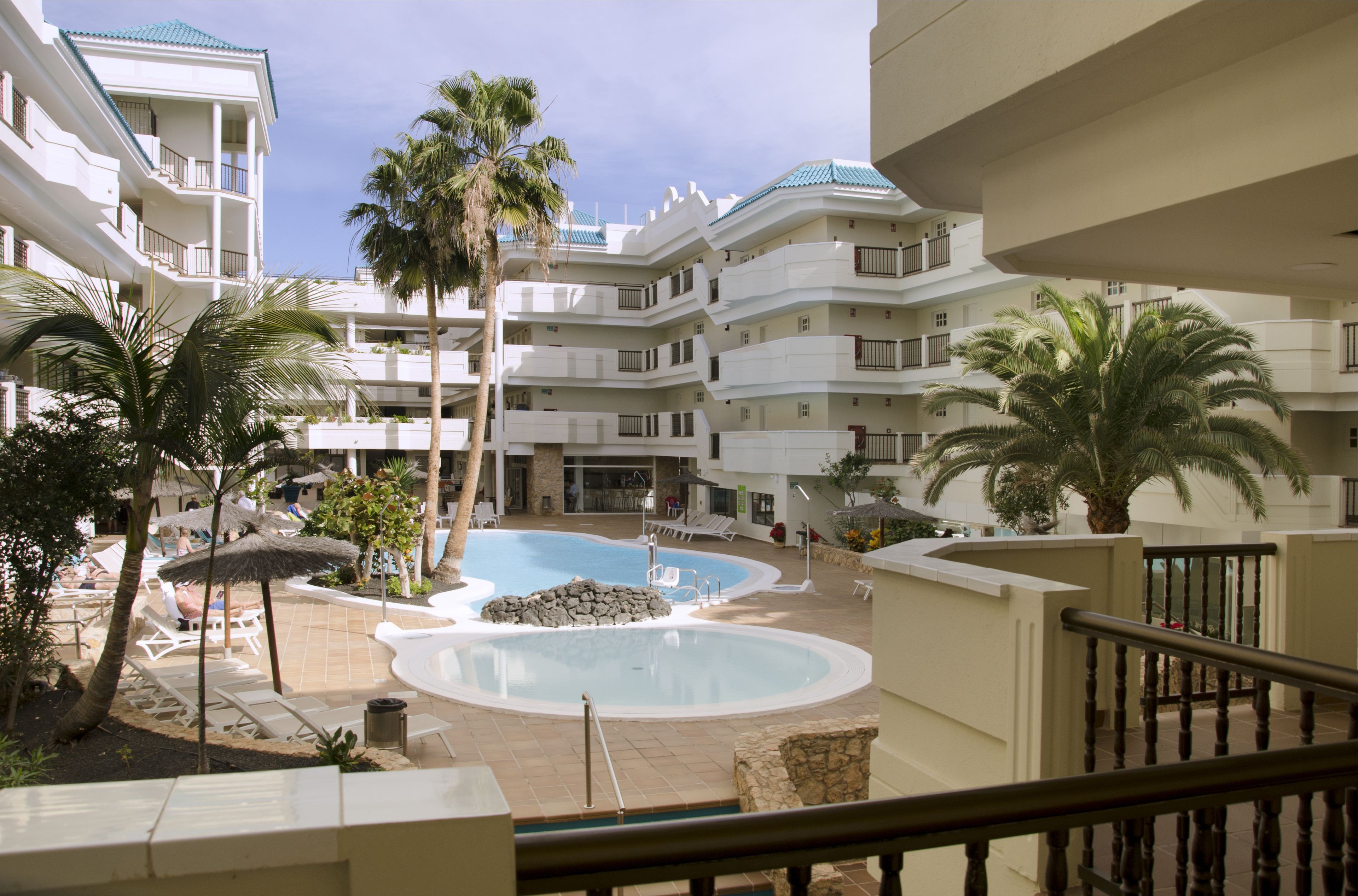A hotel with a central swimming pool, multiple balconies, and palm trees.