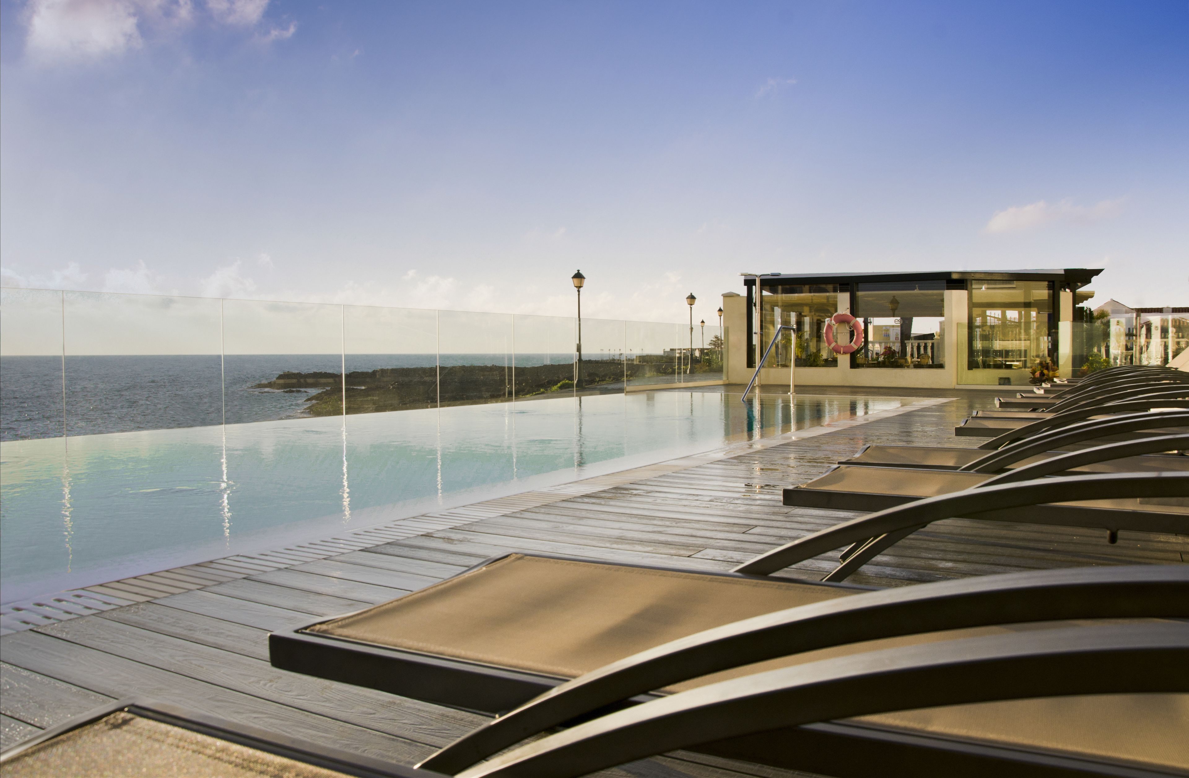 Un espace aménagé au bord de la piscine, avec des chaises longues, et un bâtiment doté d’une façade en verre donnant sur la mer.