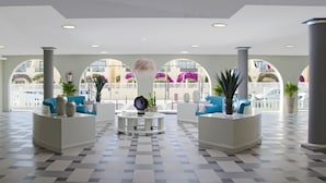 A spacious lobby with checkered flooring, two seating areas with blue sofas, a central round table, and large windows offering views of a white fence and buildings. - Hotel Ereza Mar- Adults Only (Antigua)