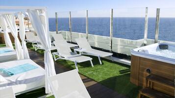 A rooftop deck with white lounge chairs, a hot tub, and a glass balustrade overlooking the sea.