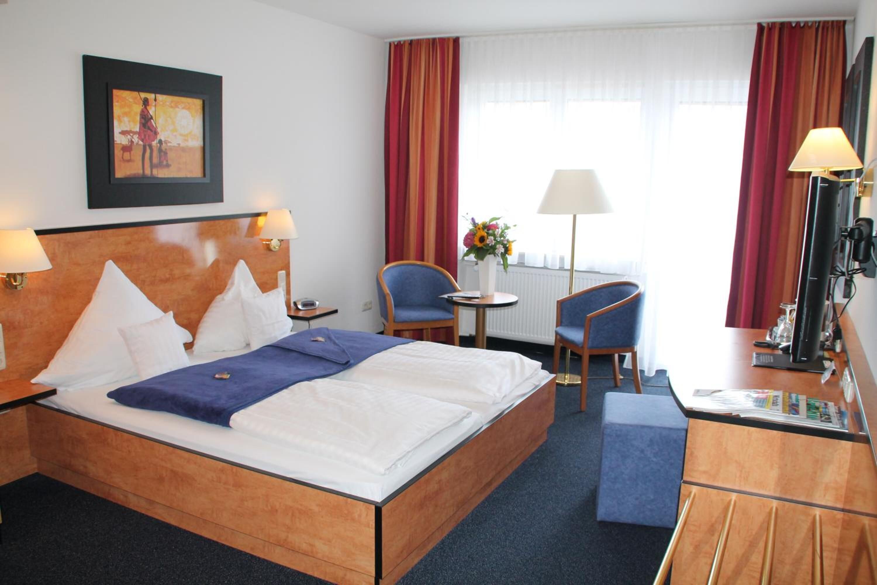 A hotel room with a large bed, two chairs, a desk with a lamp, a small table with a vase, and a picture on the wall.