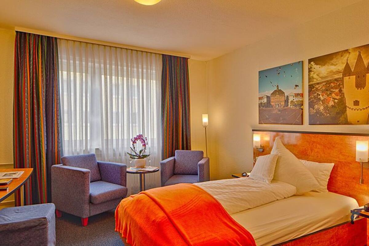 A hotel room with a bed, two armchairs, a desk, and a small table with a vase of flowers.