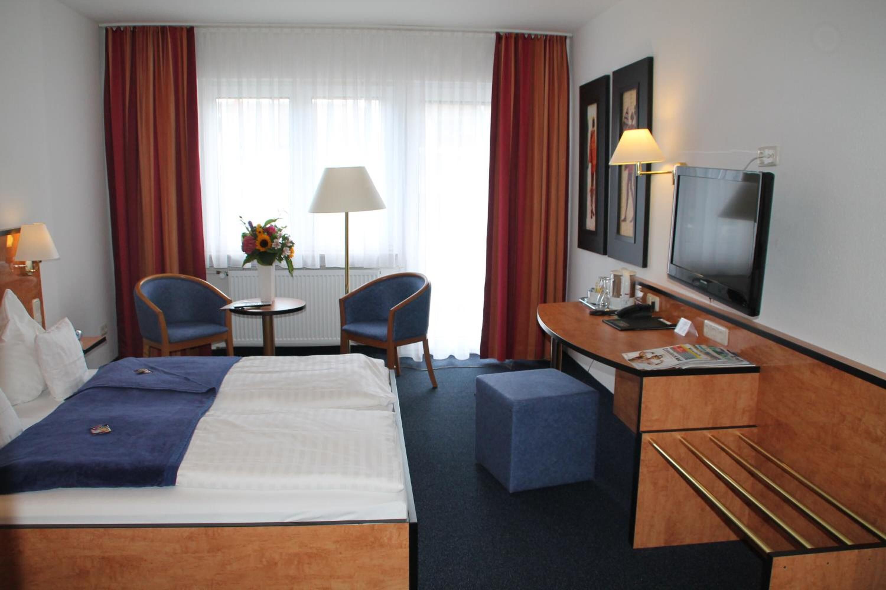 A hotel room with a bed, two chairs, a desk with a TV, a lamp, and a window with curtains.