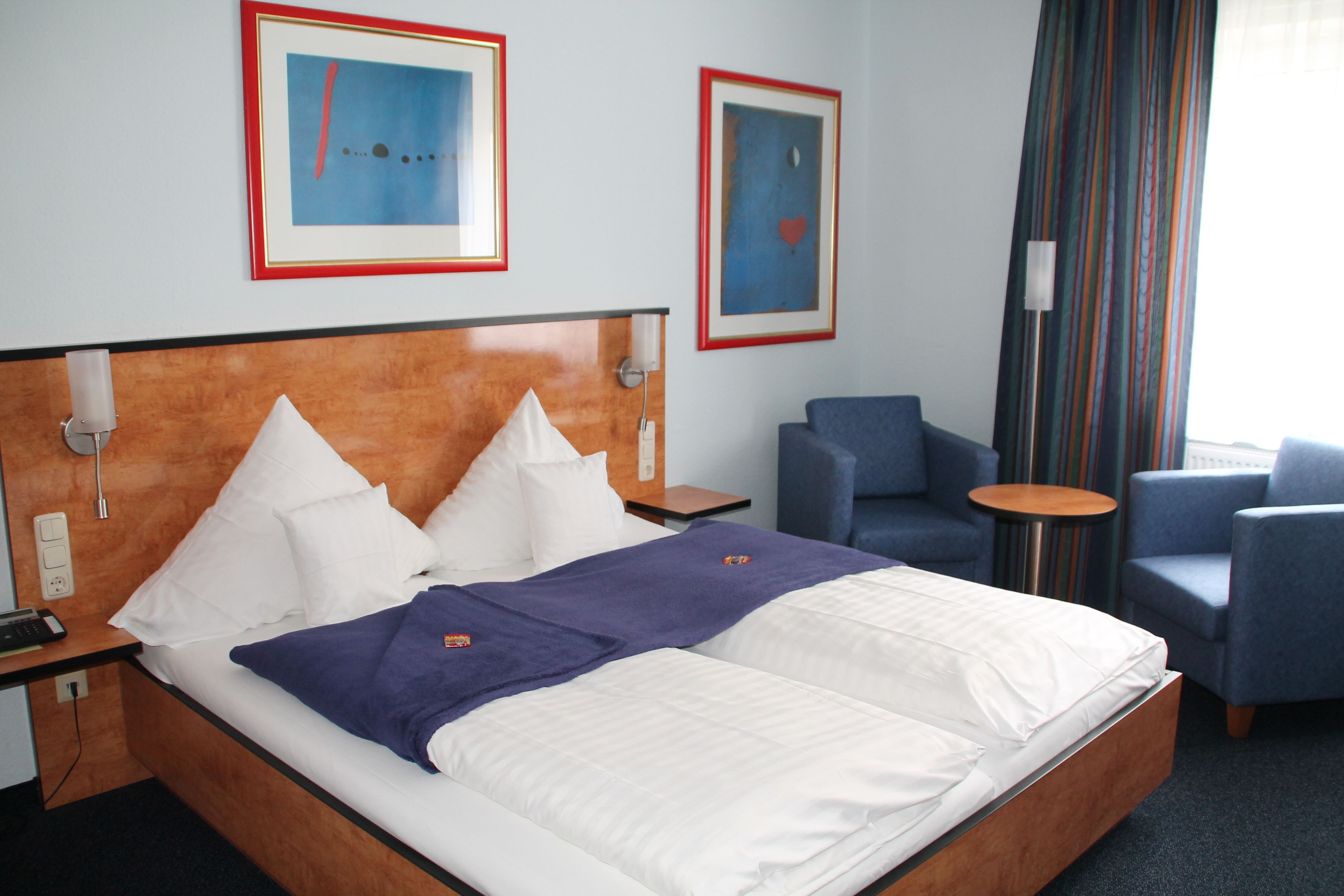 A hotel room with a large bed, two side tables, a desk, a chair, and two framed artworks on the wall.