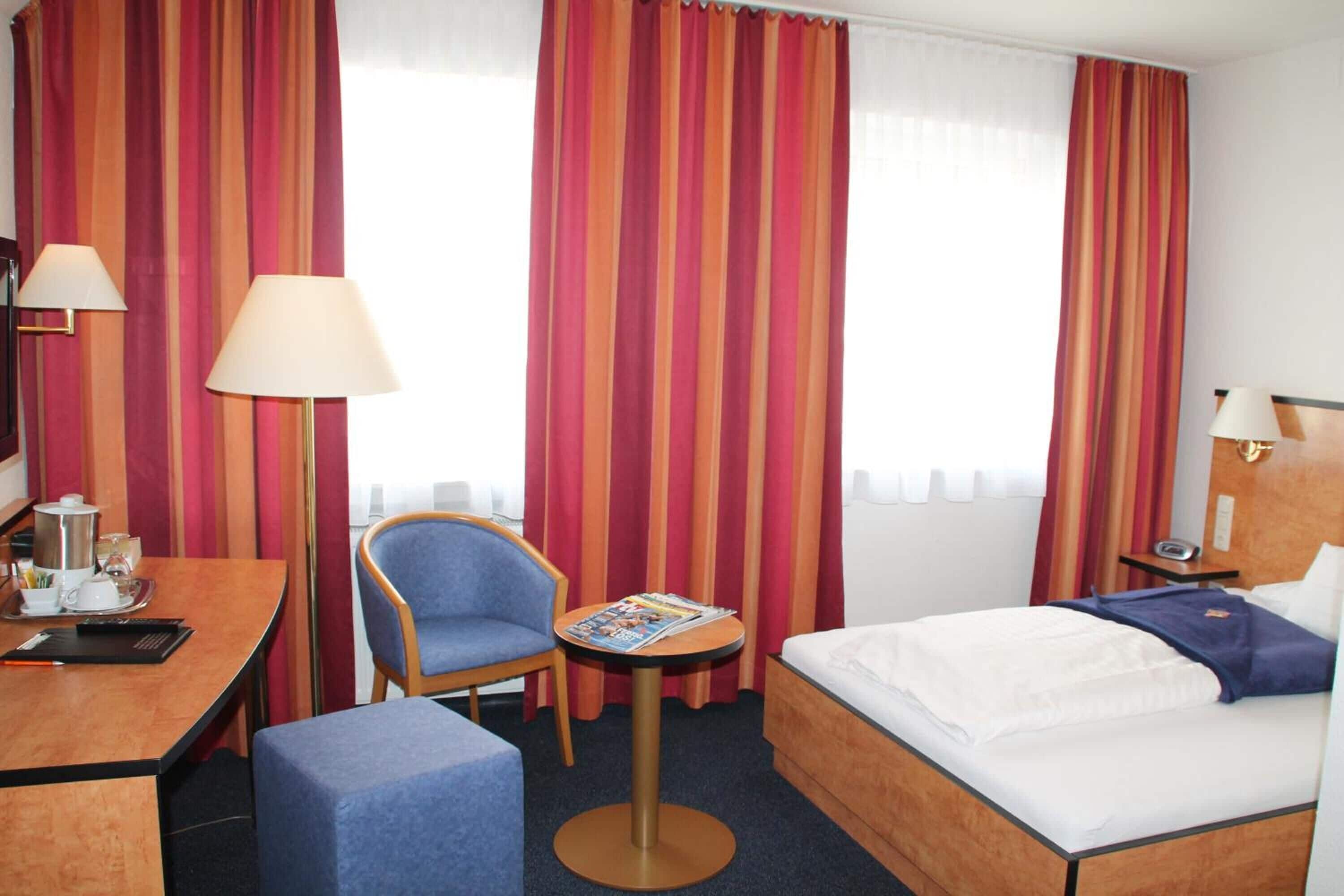 A hotel room with a bed, a desk, a chair, a small table, and curtains.