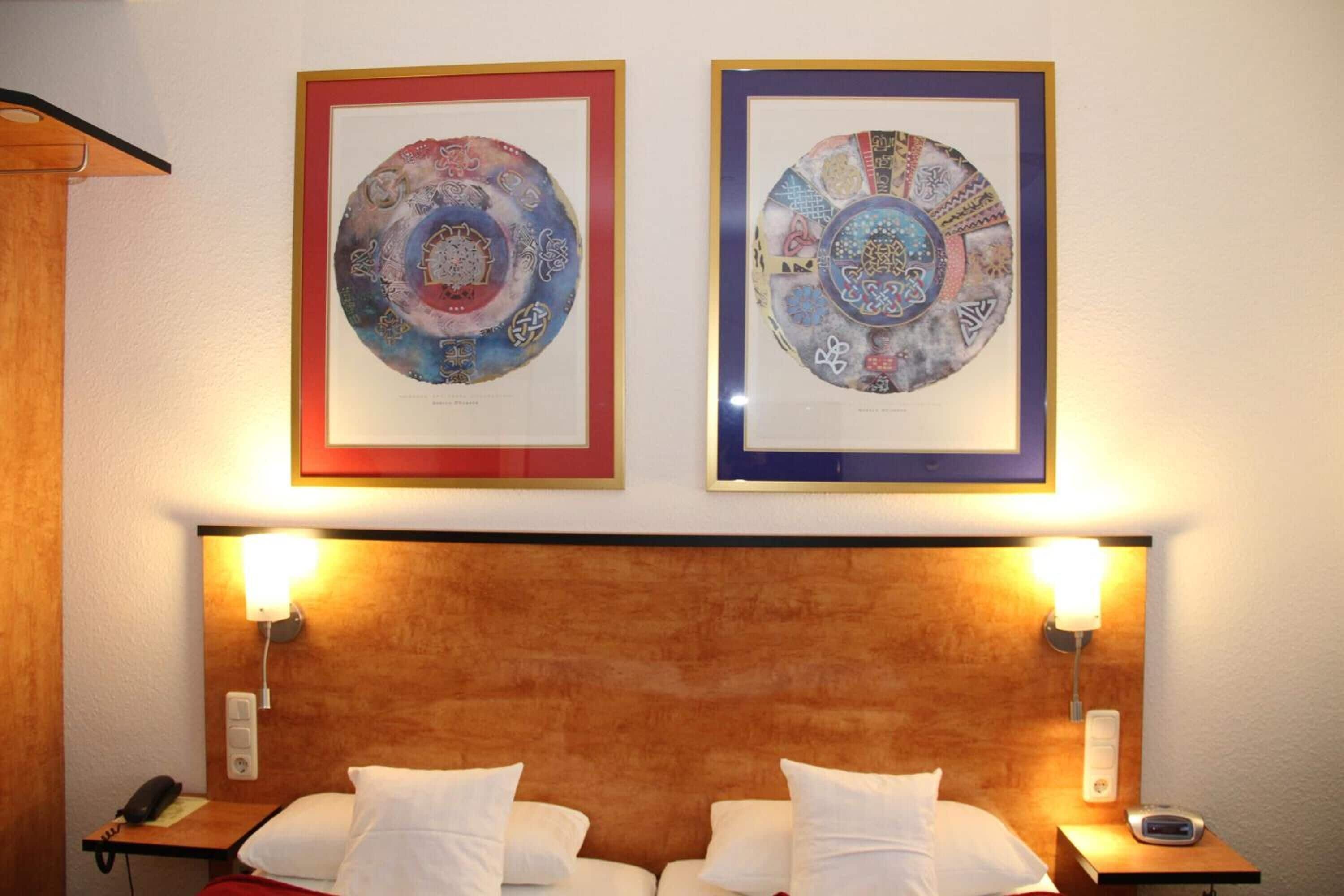 Une chambre d’hôtel avec deux lits, une tête de lit en bois, des œuvres d’art encadrées au-dessus des lits, des lampes de chevet et un téléphone sur la table de chevet.