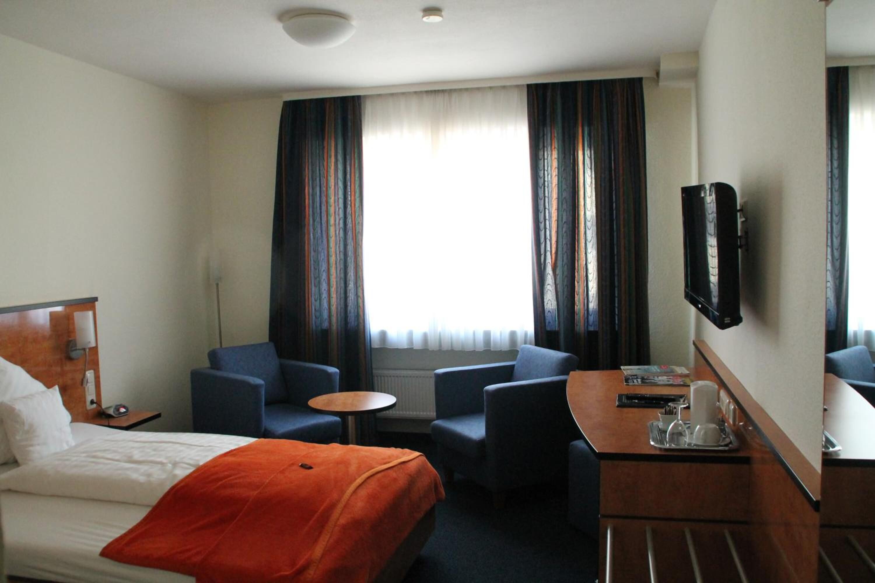 Ein Hotelzimmer mit einem Bett, zwei Sesseln, einem kleinen Tisch, einem Fernseher und einem Fenster mit Vorhängen.