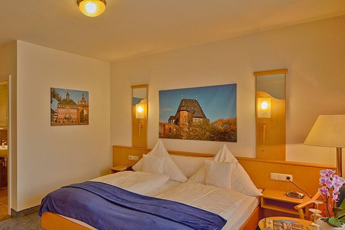 A hotel room with a bed, a desk, a chair, a lamp, a mirror, and two paintings on the wall.