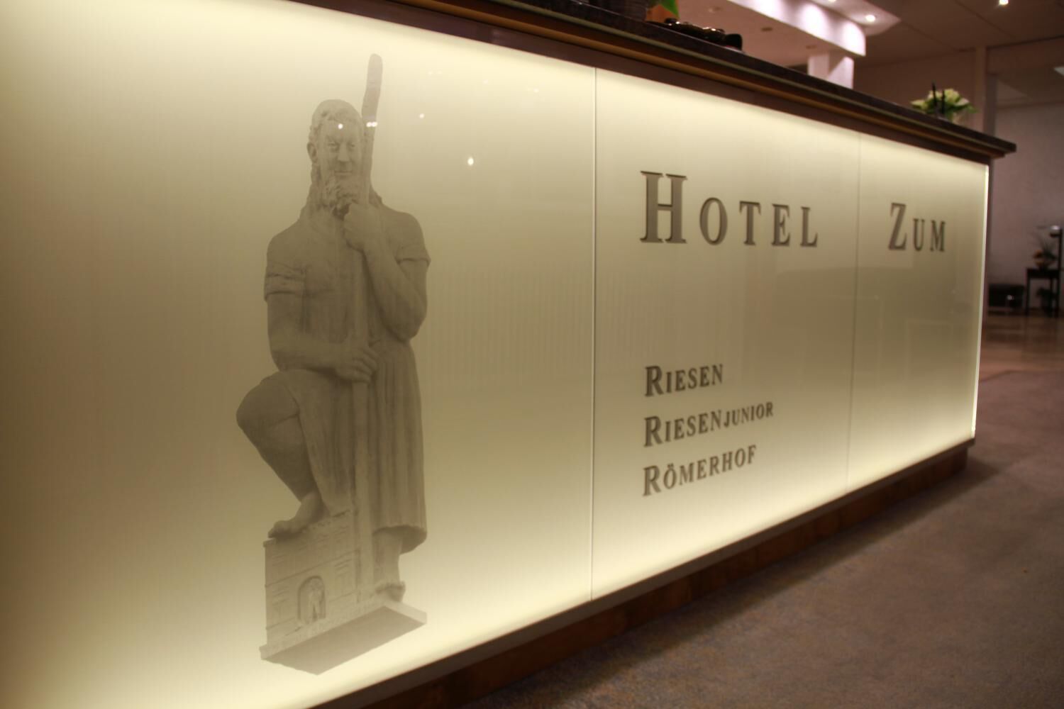 A reception desk with a statue of a man holding a staff, and the text "Hotel Zum Riesen Riesenjunior R\"omerhof" is visible.