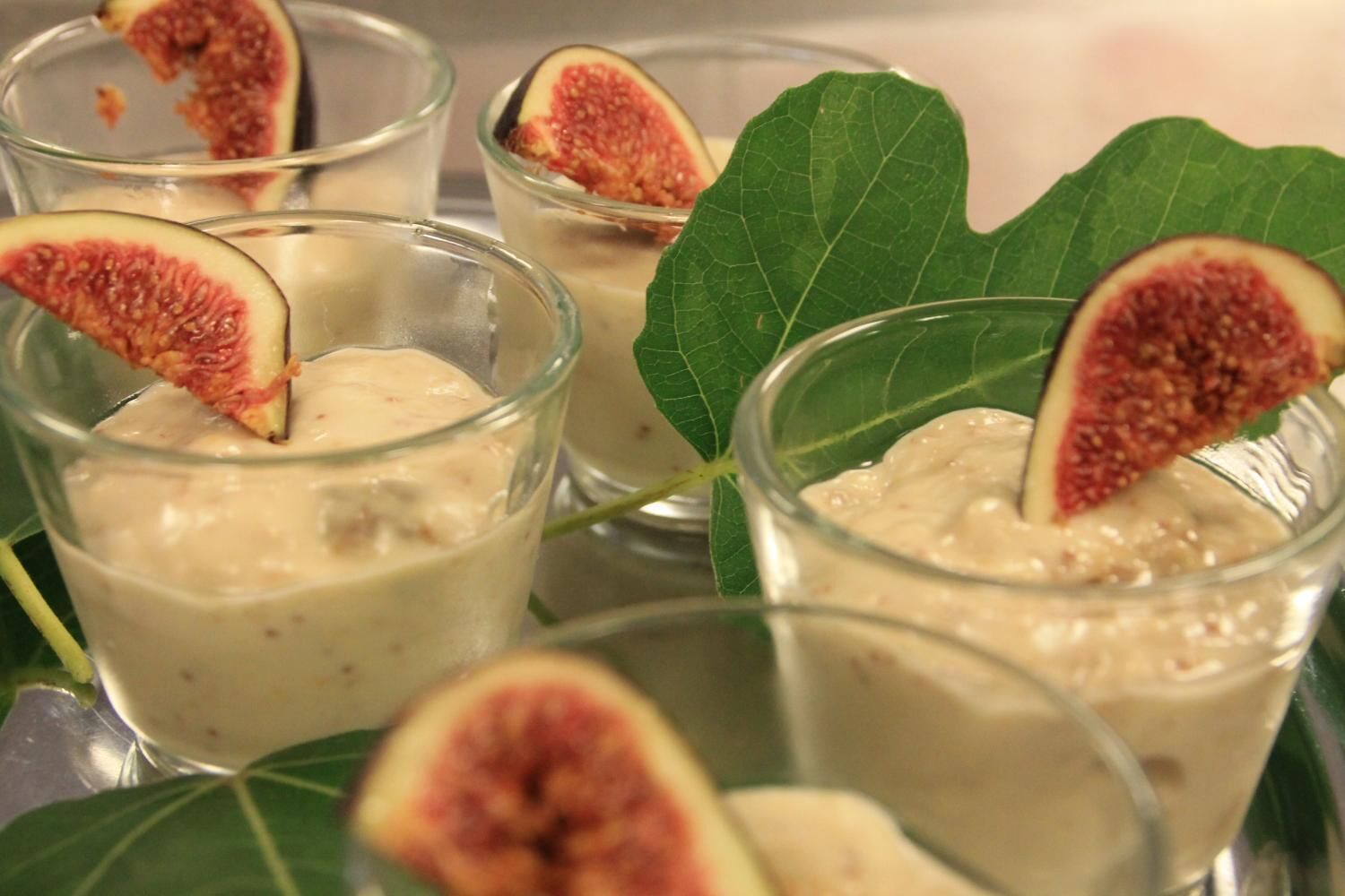 Quatre verres contenant un dessert crémeux aux figues, servis sur une surface réfléchissante et décorés d’une feuille verte.