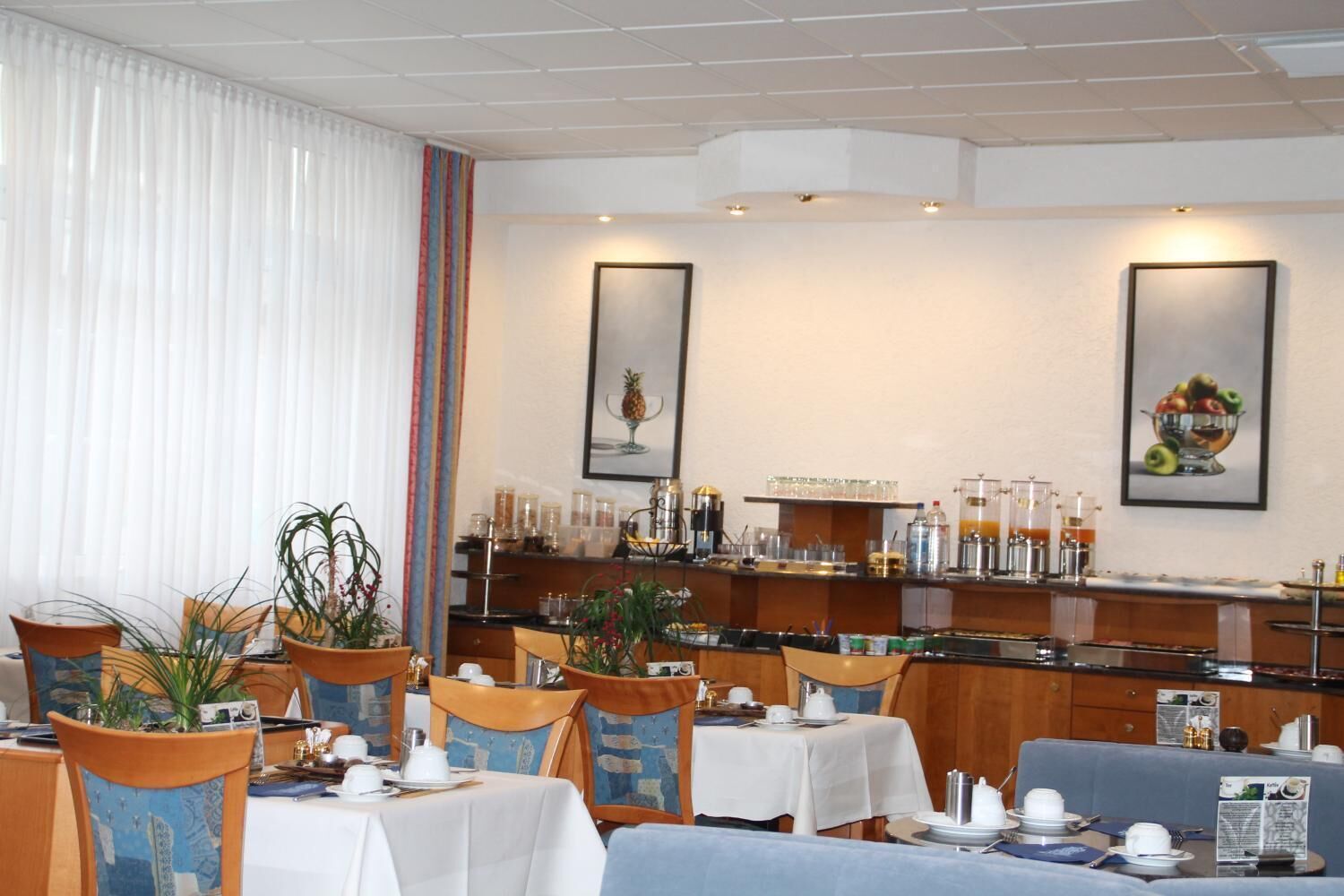 A hotel dining area with tables set for breakfast, a buffet counter with various food items, and two framed pictures on the wall.