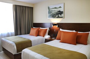 Habitación de hotel con dos camas, cada una con colcha marrón y blanca y almohadas naranjas. Hay una mesita de noche con lámpara entre las camas. Un cuadro de un paisaje costero cuelga en la pared sobre las camas.