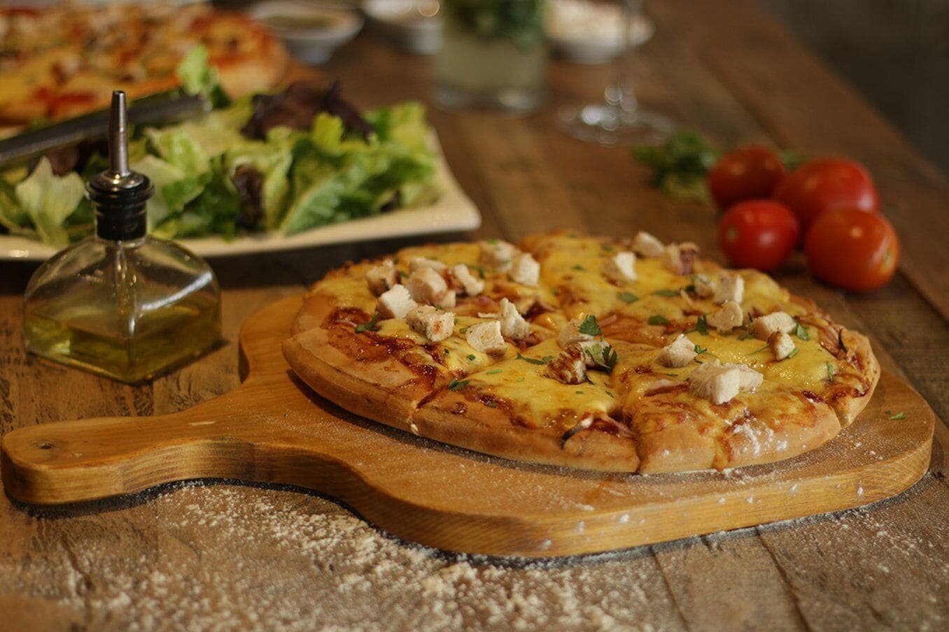 A pizza on a wooden board with a bottle of oil and some tomatoes in the background.