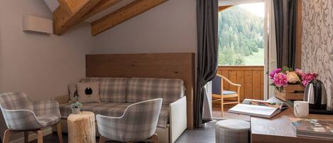 A cozy room with a wooden ceiling, a plaid sofa, two chairs, a small wooden table, and a view of a forest through a window.