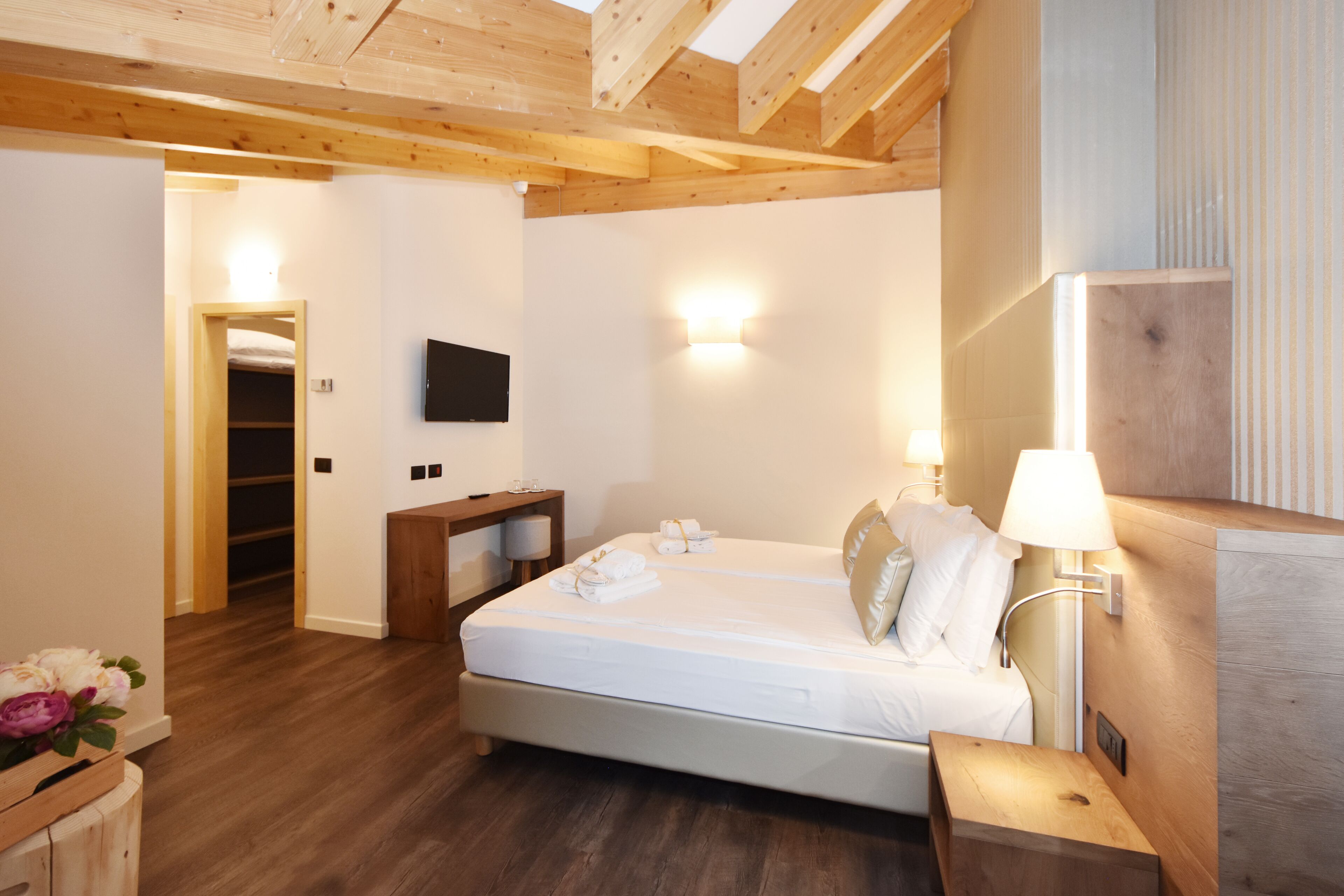 A hotel room with a wooden ceiling, a bed with white linens, a bedside table with a lamp, and a TV mounted on the wall.