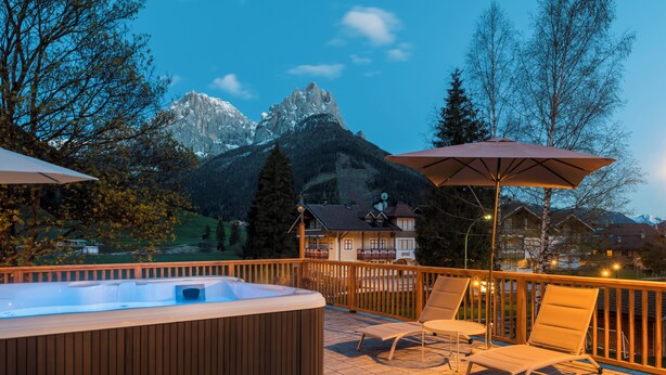 A wooden deck with a hot tub, lounge chairs, and umbrellas, overlooking a mountain and a village.