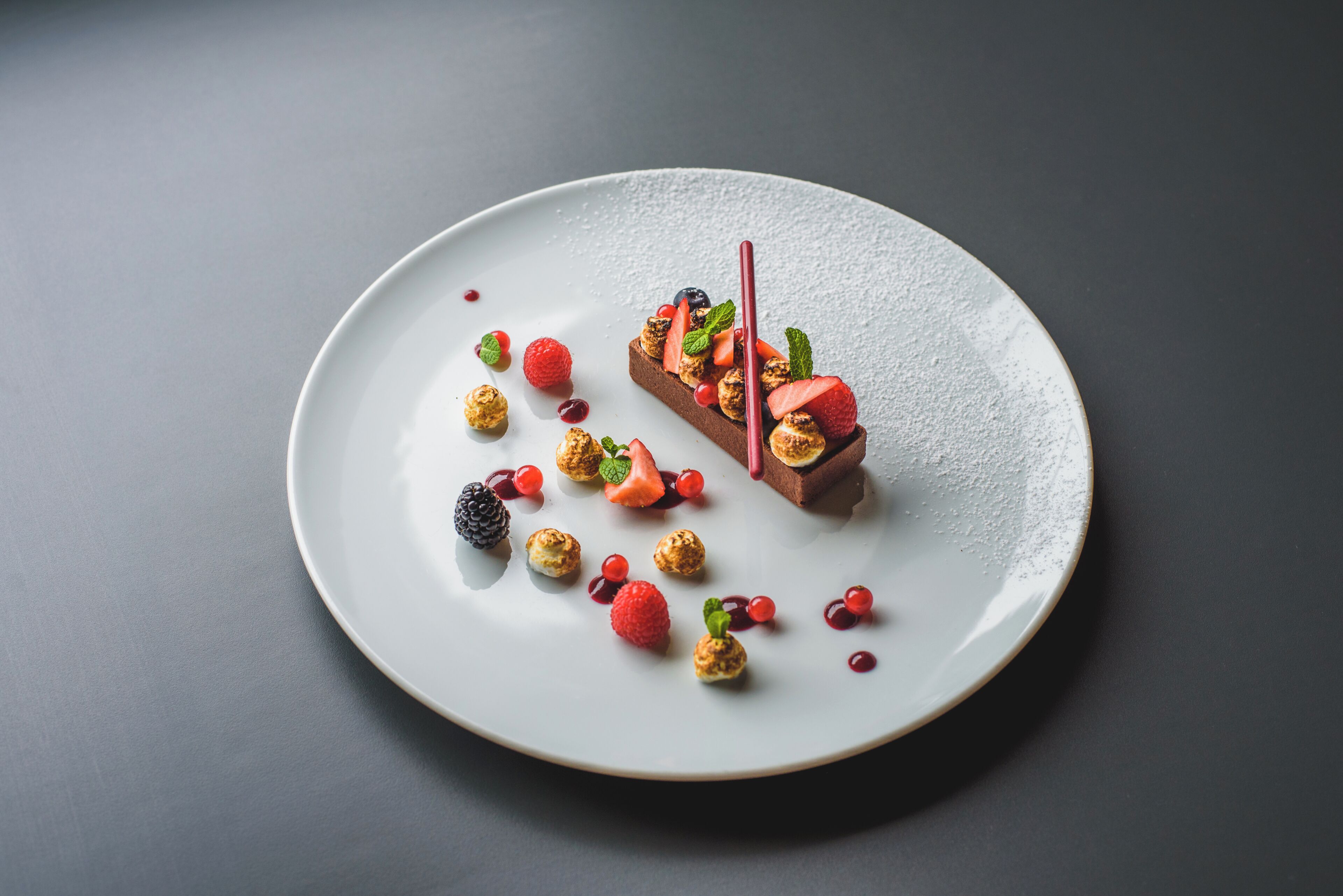 Un postre en un plato blanco con bayas y un trozo de chocolate.